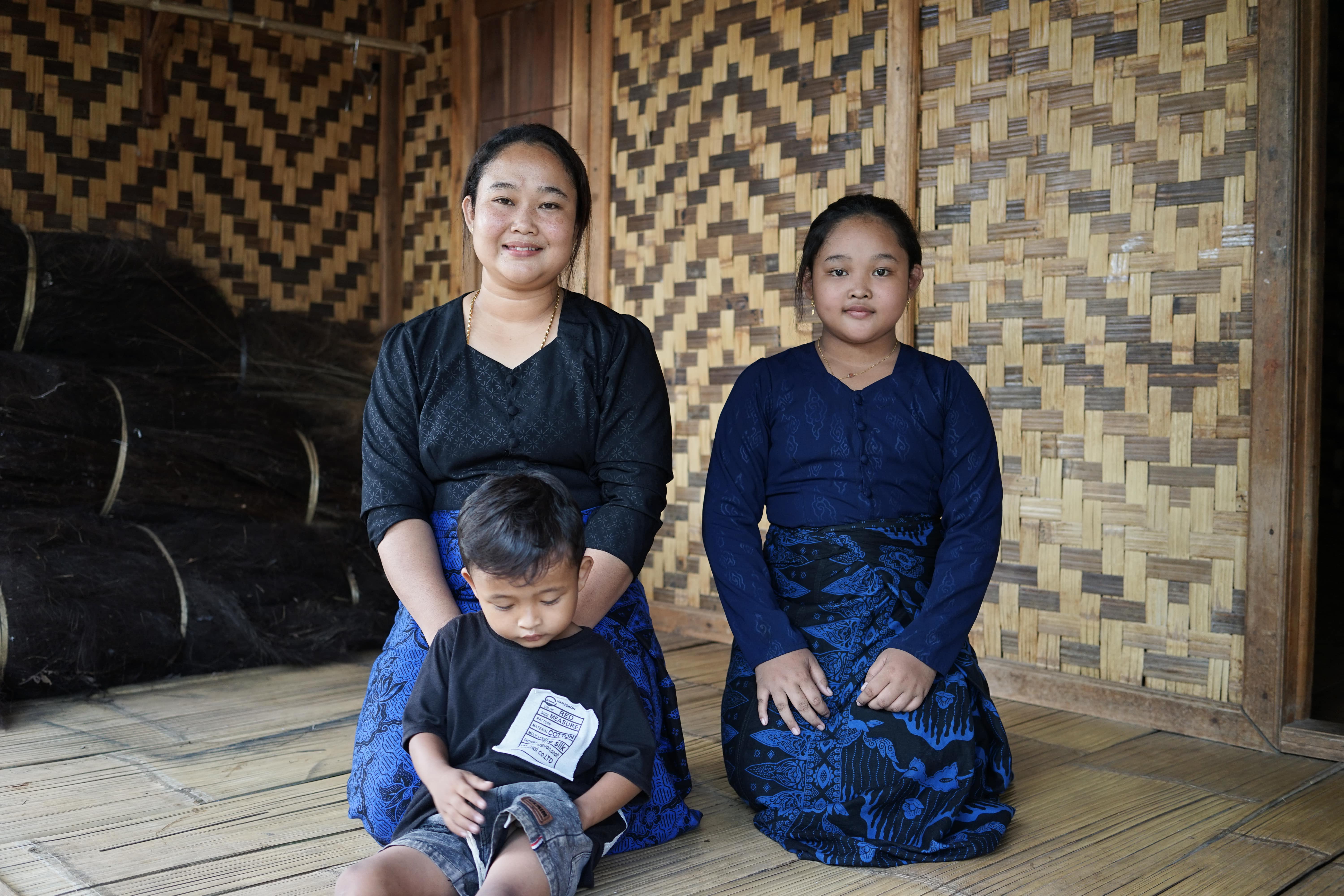 A mother, her daughter, and her young son sitting on a wooden floor against a backdrop of woven bamboo walls.