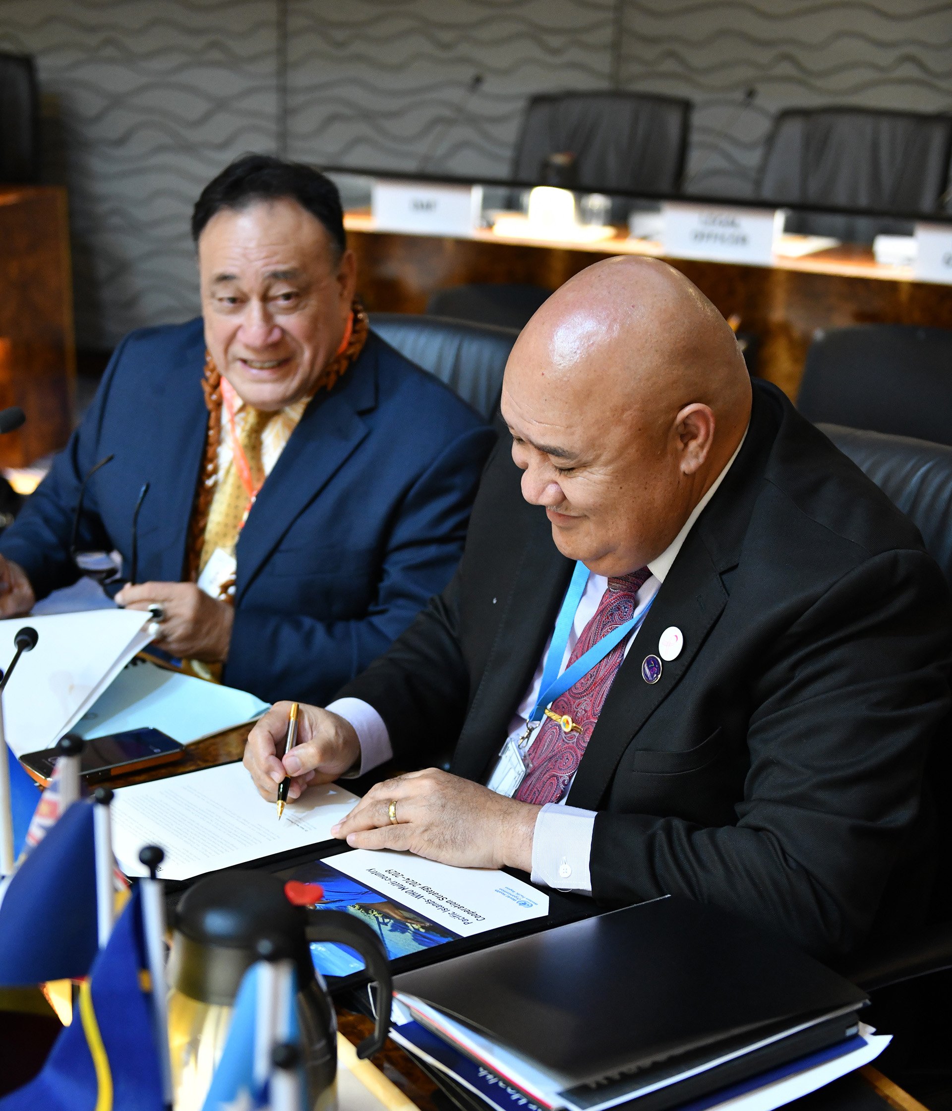 Close-up of two individuals at a formal international event, seated at a table. One is writing on a document. The setting is a professional conference with nameplates and several small flags.